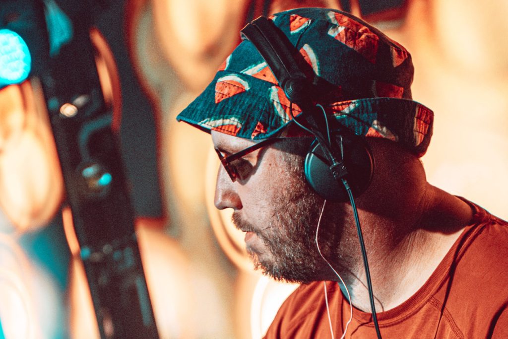 Close-up profile of a Dub Catalyst band member in headphones and a watermelon-print bucket hat, performing live at Mucky Weekender 2026.